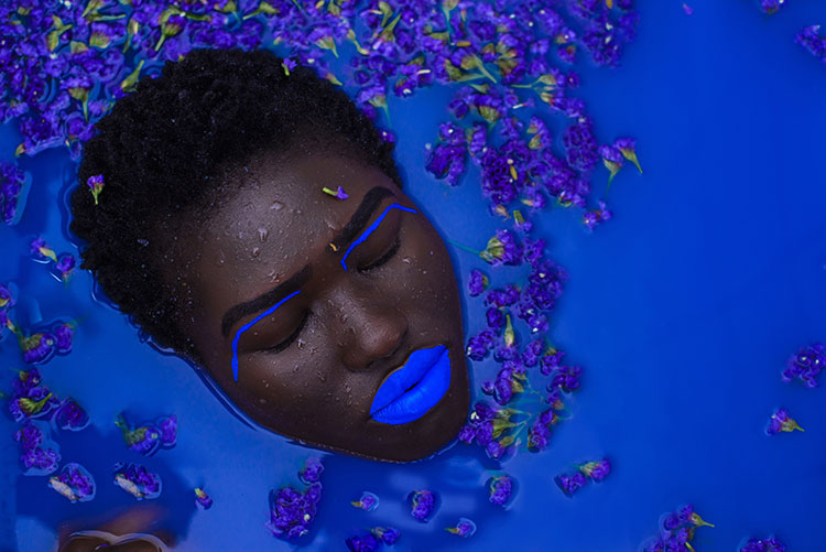 Br24 Blog Visuelle Trends 2020: Trend - Makeup is Not a Mask; Porträt einer dunkelhäutigen Frau mit blau geschminkten Lippen und blauem Augen Makeup, nur ihr Kopf ist zu sehen wie sie in einer dunkelblauen Flüssigkeit schwimmt