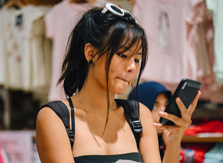 Br24 Blog Gen Z Shopping: Young woman inside a clothing shop, looks sceptically at her mobile phone